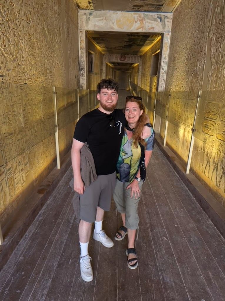 A smiling man and woman stand in an ancient Egyptian tomb corridor, its hieroglyphic walls protected by glass barriers.