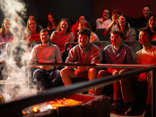 An audience seated in a dark theater watches molten lava being poured into a trench, glowing yellow-orange under stage lights.