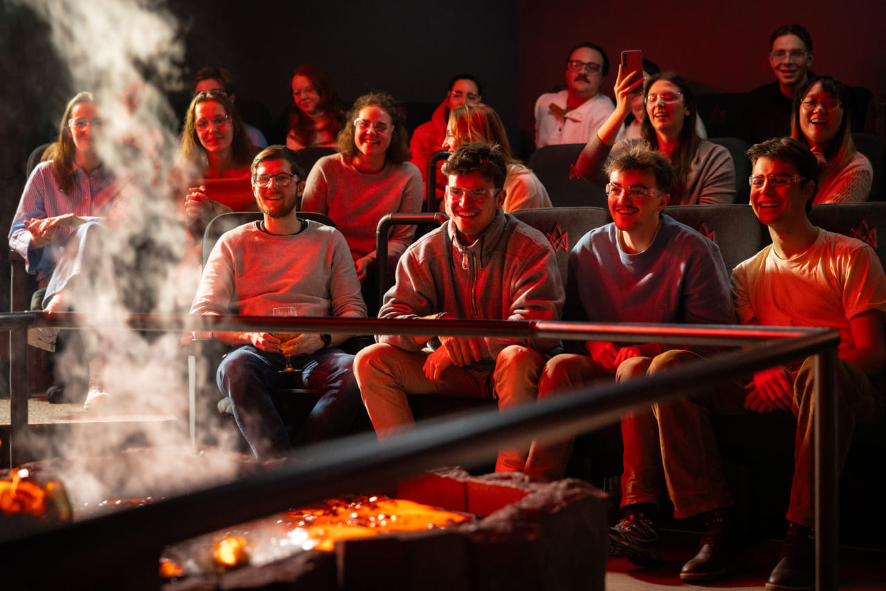 An audience seated in a dark theater watches molten lava being poured into a trench, glowing yellow-orange under stage lights.