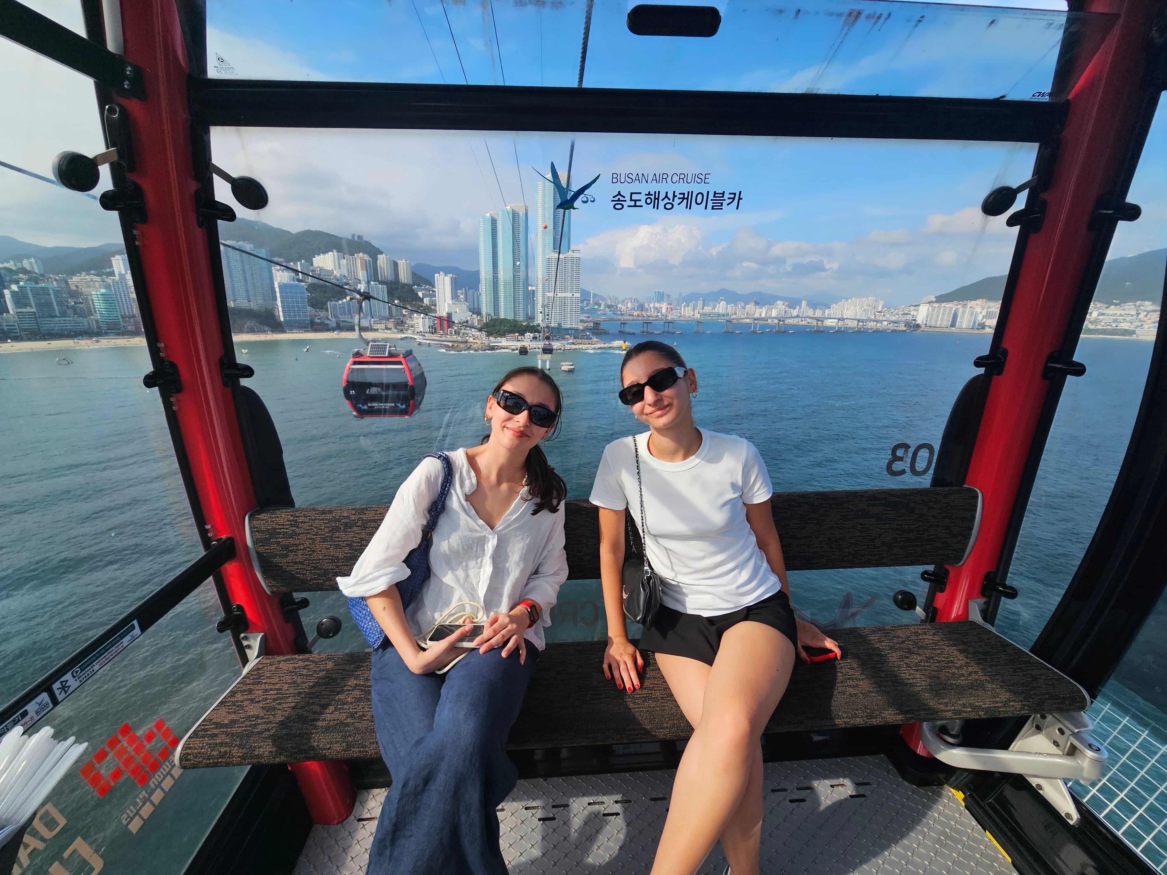 Two friends sit inside a Songdo Marine Cable Car gondola, with Busan's coastline and skyline visible through the windows.