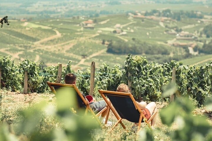 Two people are relaxing in folding lounge chairs. They are sat in the middle of a vineyard and looking at spectacular countryside landscape