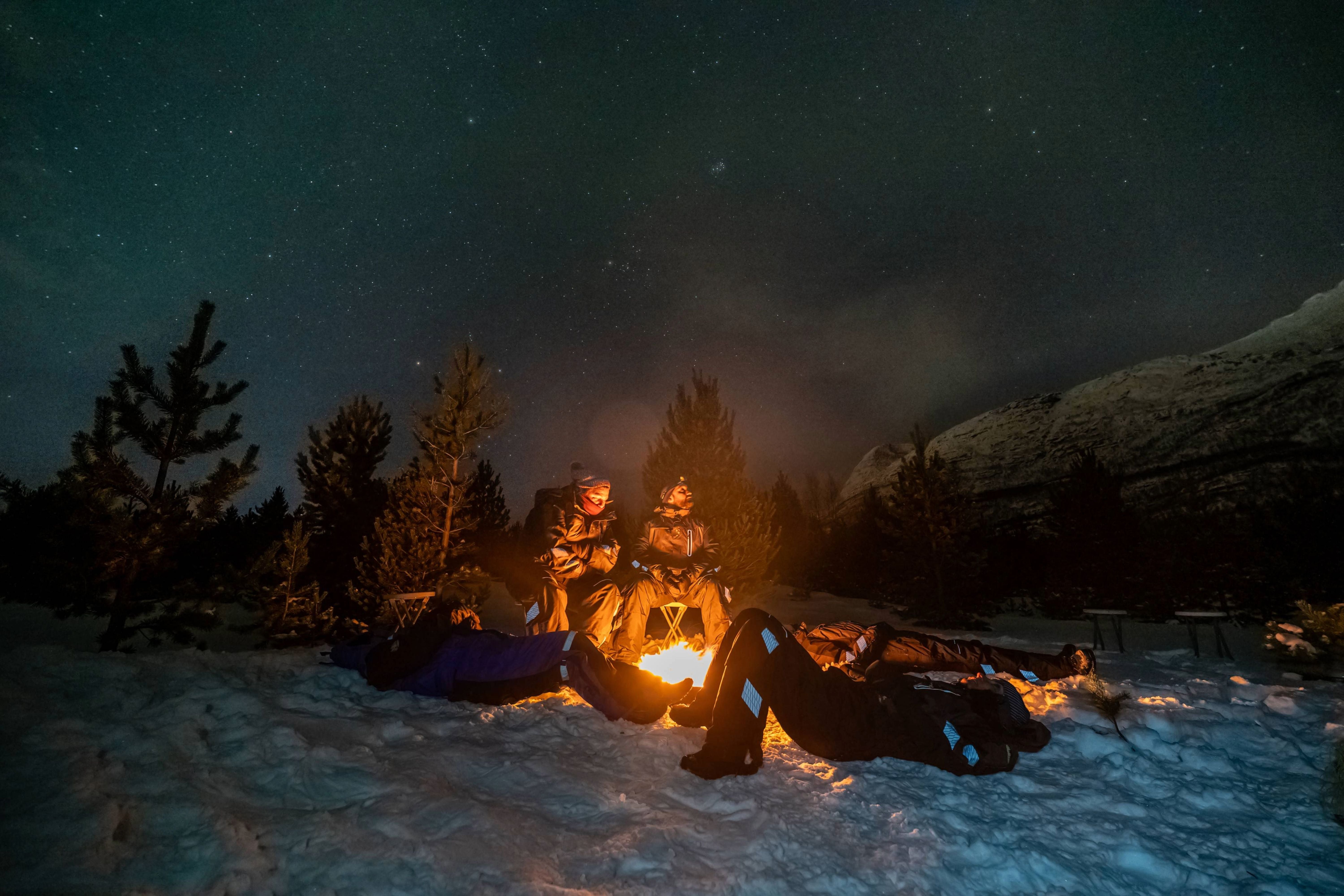 Travelers in warm thermal suits relaxing in the snow around a glowing campfire during a private Tromsø Northern Lights tour.