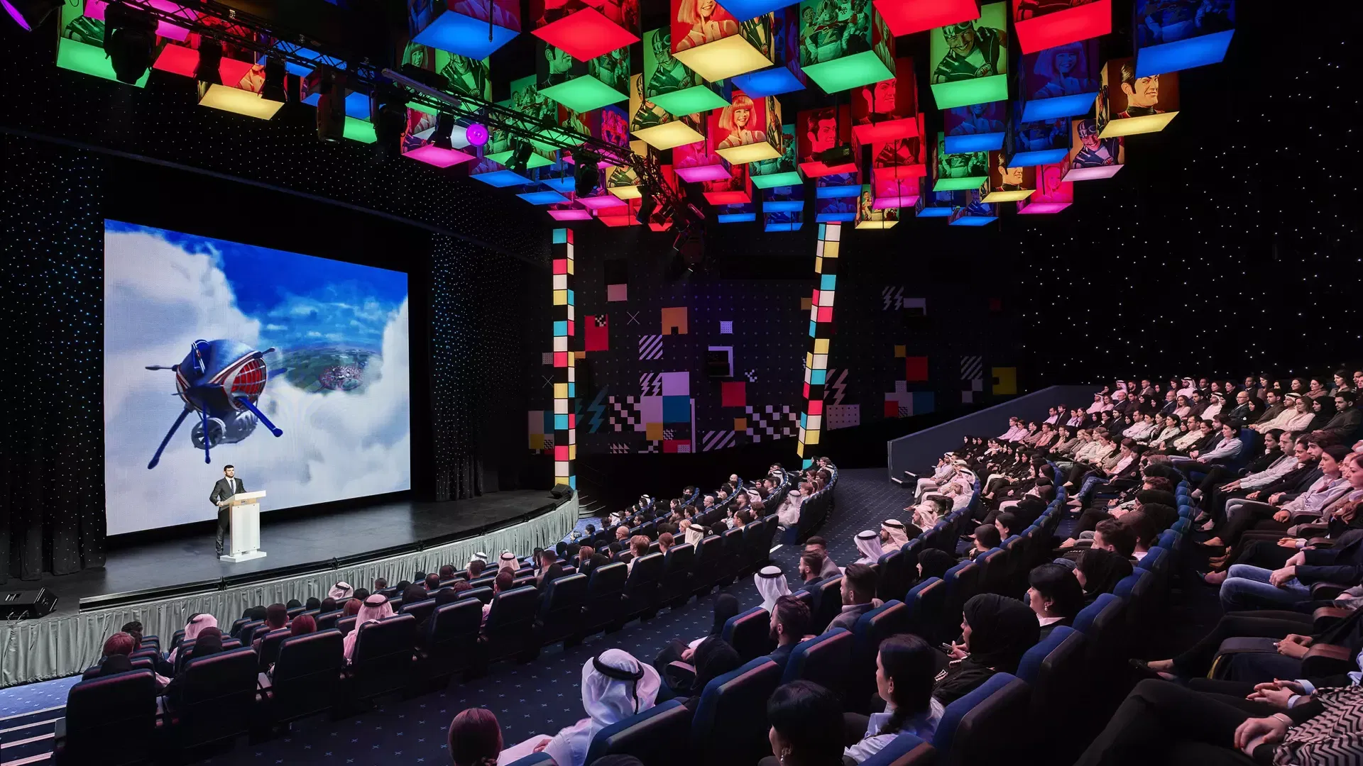 A large indoor theater features a wide presentation screen showing an animated airship flying over clouds, with an audience seated in blue chairs and colorful illuminated cubes suspended from the ceiling at IMG Worlds of Adventure Theme Park.