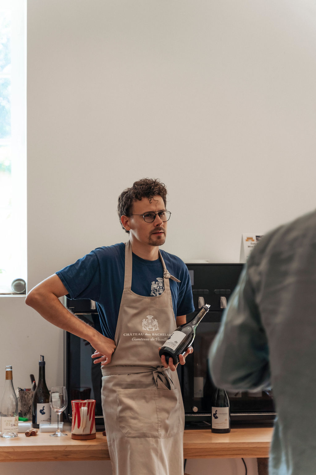 A man in his early thirties, european with brown hair, glasses and facial hair, wearing an apron with a crest and cradling a bottle of wine