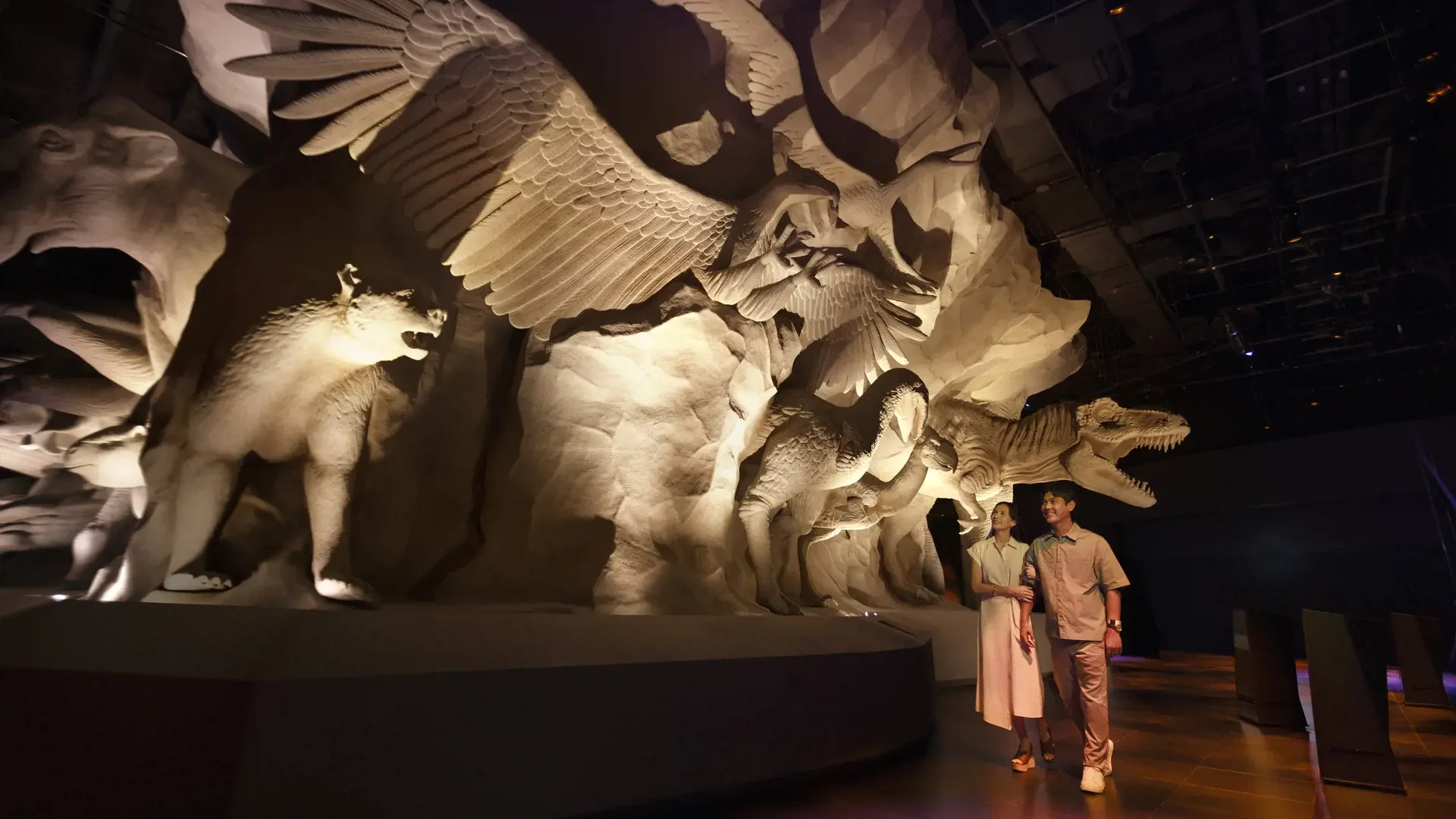 A man and a woman walk past a large, textured wall relief depicting prehistoric animals and winged creatures at Exploria in Mandai Singapore Ticket.