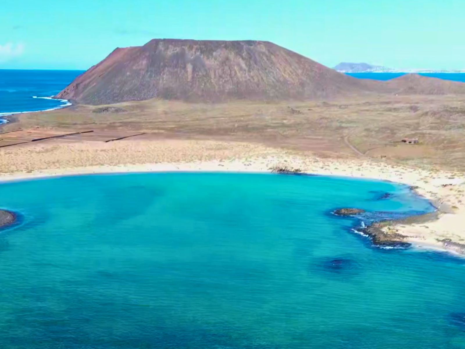 Vista aérea de la Playa de La Concha de Lobos destacando sus tranquilas aguas turquesas, arenas doradas y paisajes volcánicos.