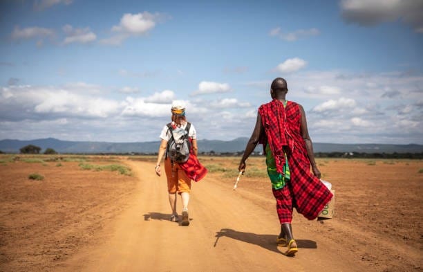 Rear view of a tourist with a backpack walking down a dirt road next to a Maasai host wearing a red shúkà under a clear blue sky.