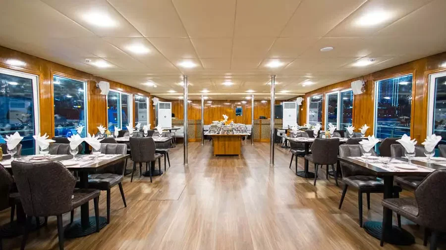 Inside the indoor dining area, rows of dark cushioned chairs and tables are set with white napkins, beneath a tiled ceiling and large windows at Alexandra Sea Lounge.