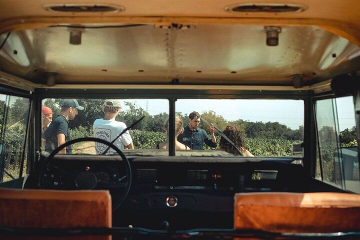 View from inside a vintage Land Rover, looking out through the windshield. A group of people are standing in a vineyard on a sunny day.