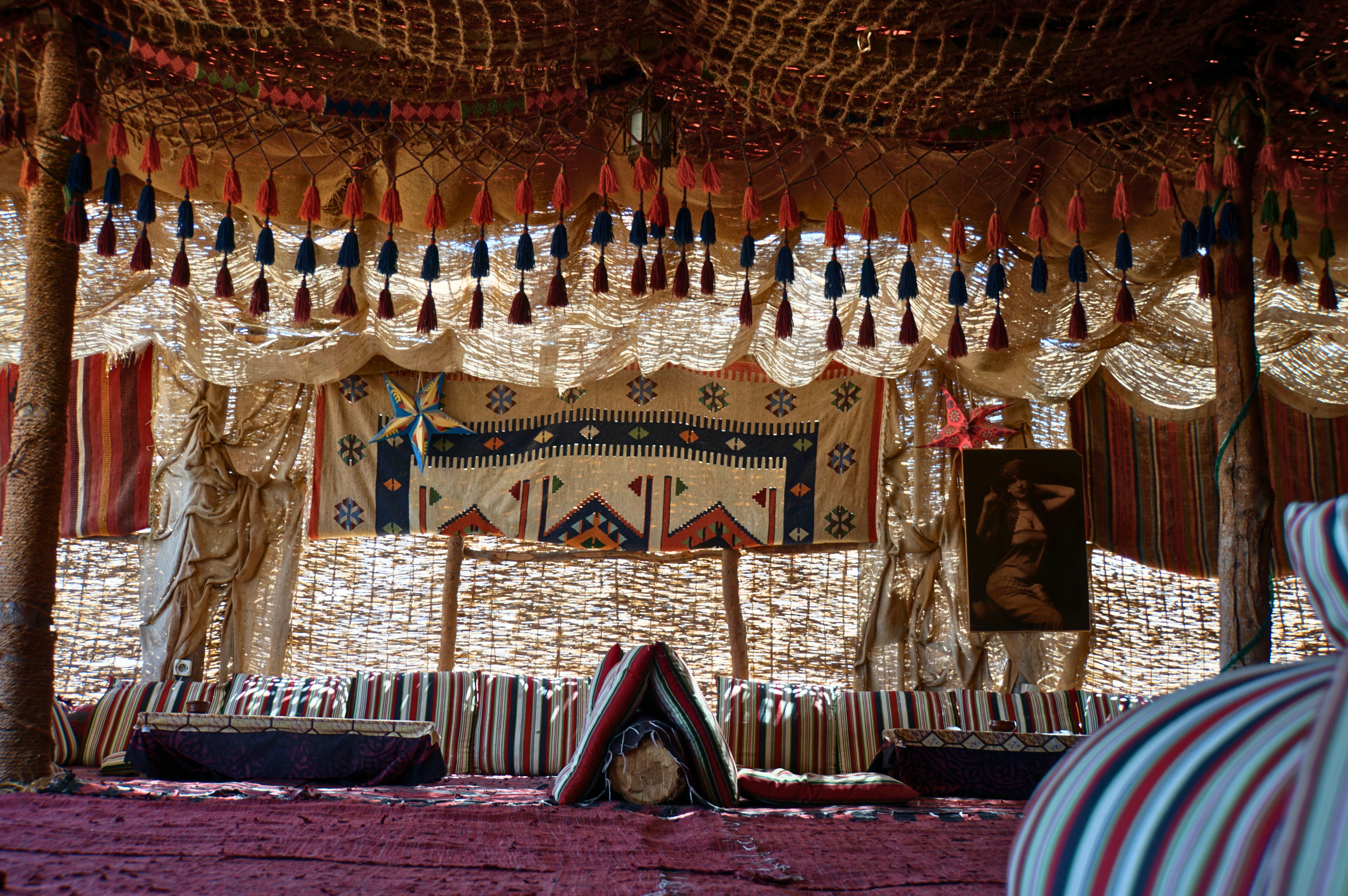 Interior view of a temporary Bedouin rest tent with patterned carpets, striped cushions, tassels, and traditional woven décor.