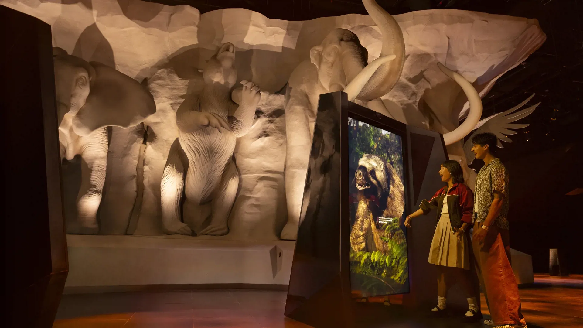 Two people stand before an interactive screen displaying an animal, with large white stone animal sculptures mounted on the wall behind them at Exploria in Mandai Singapore Ticket.