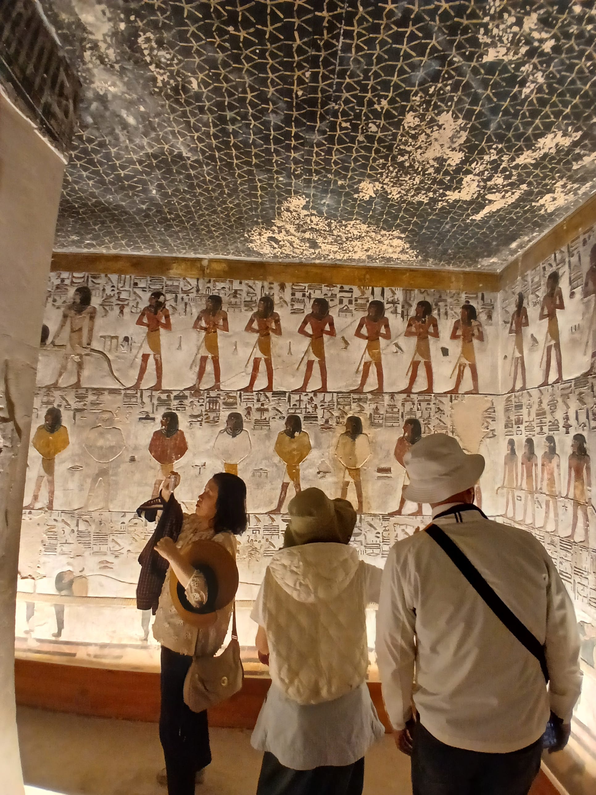 Tourists viewing a stone wall painted with yellow-background rows of ancient Egyptian deities 