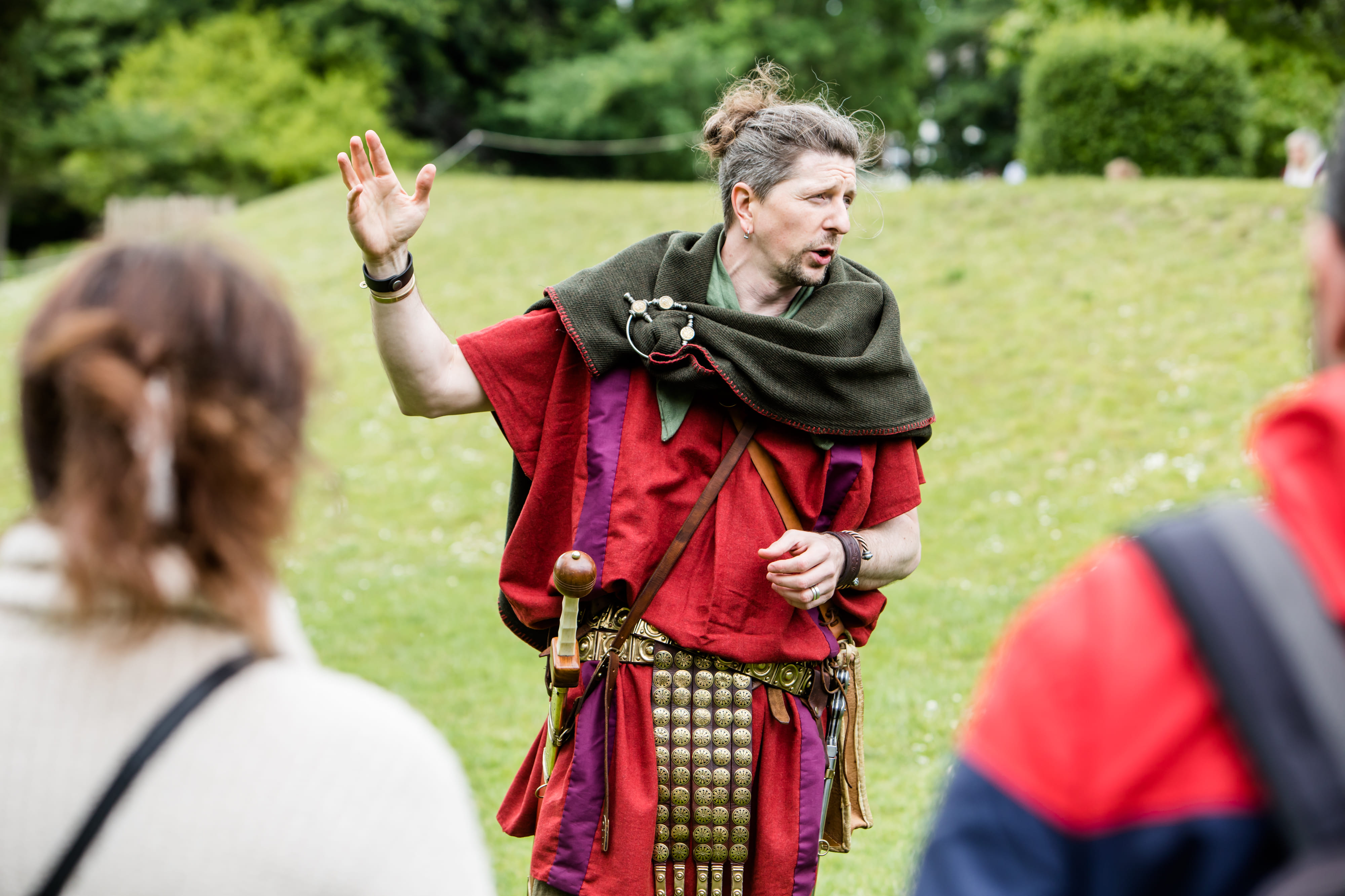 The Original Roman Walk's costumed guide is stood surrounded by guests in a lush green area of the Museum Gardens, York.