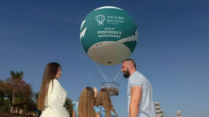 Two women, a man, and two girls stand outside looking up at a large branded tethered balloon against a clear blue sky at The Dubai Balloon at Atlantis.