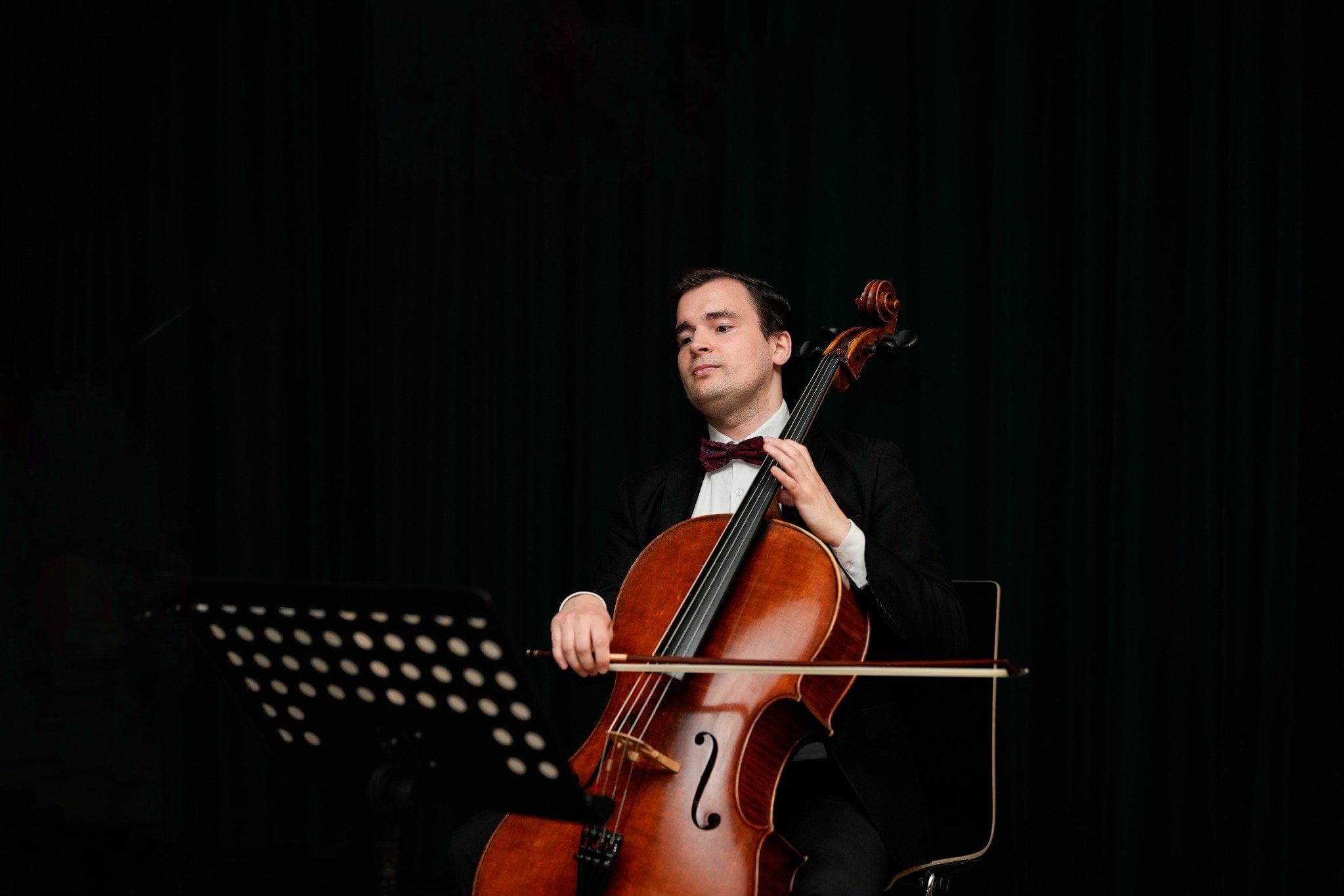 Man in a tuxedo playing cello at Haus der Musik, Vienna, seated beside a music stand.