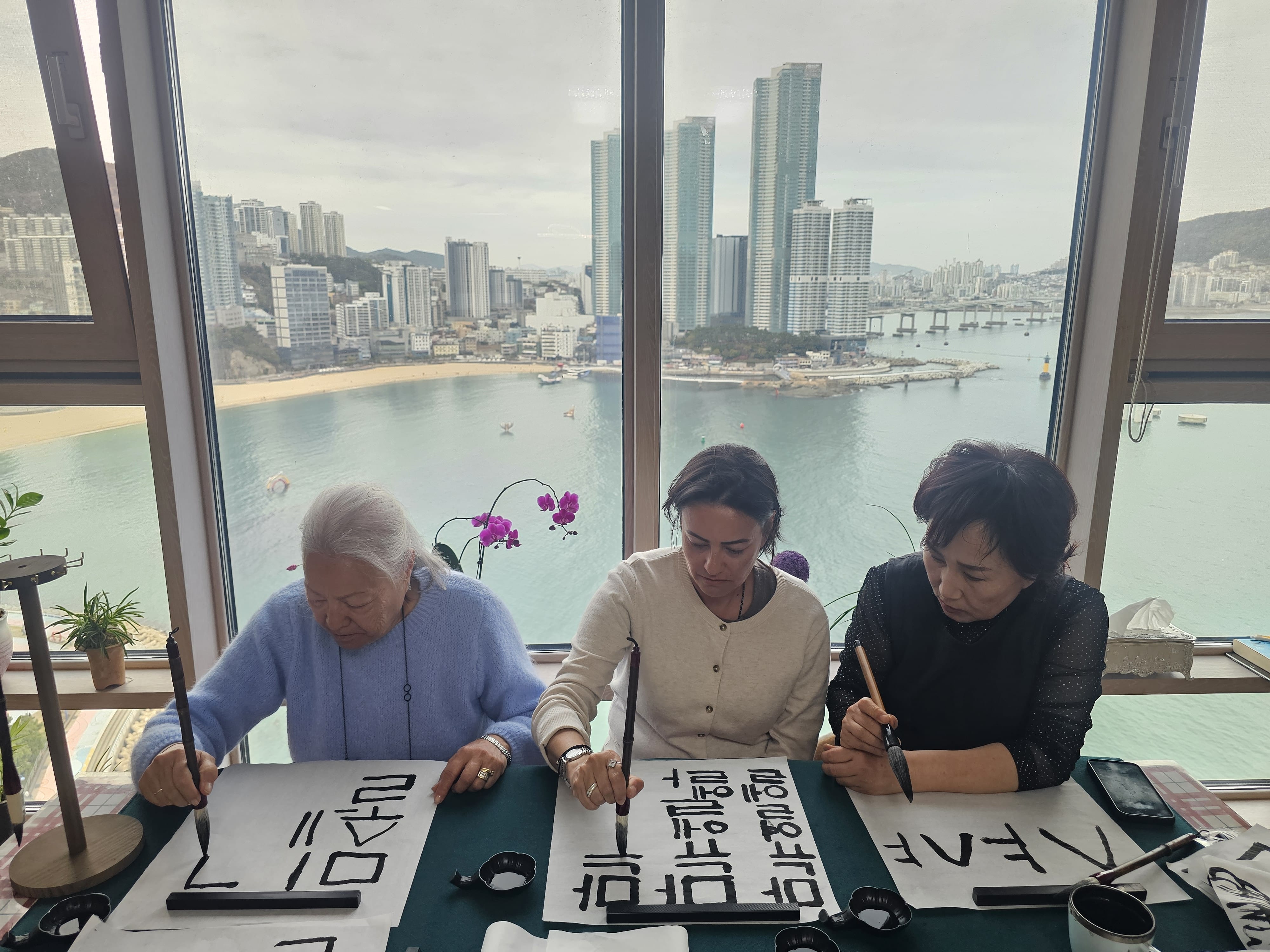 Guests practicing Korean calligraphy by writing their names with a brush inside a home, enjoying a cultural experience in Busan