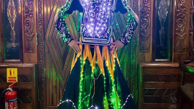 A person stands wearing a traditional outfit covered in glowing string lights against a dark wooden background at Alexandra Dhow Cruise in Dubai Marina.