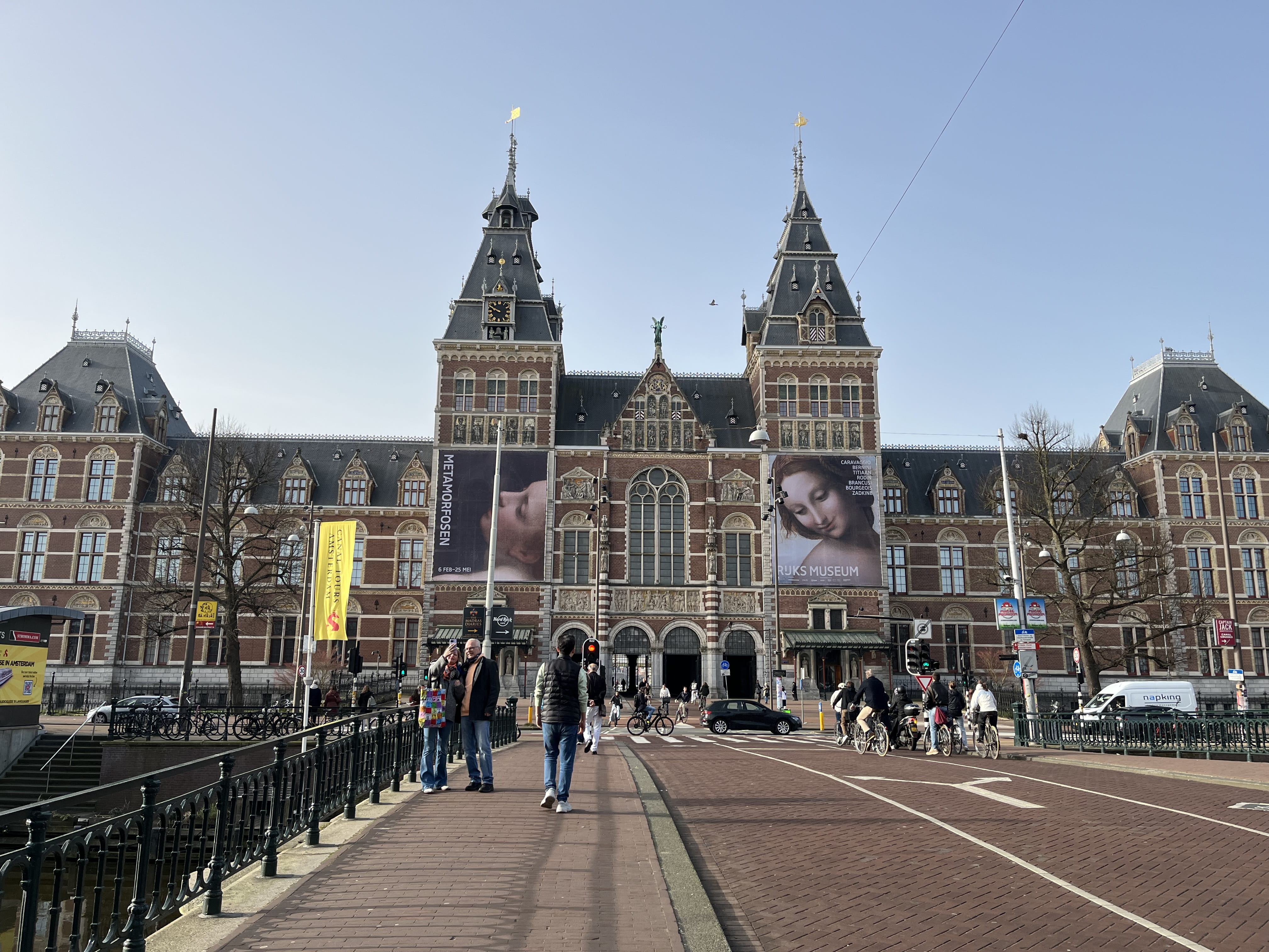 The Rijksmuseum in Amsterdam seen from a street and bridge perspective, with people walking and cycling toward the large historic building 