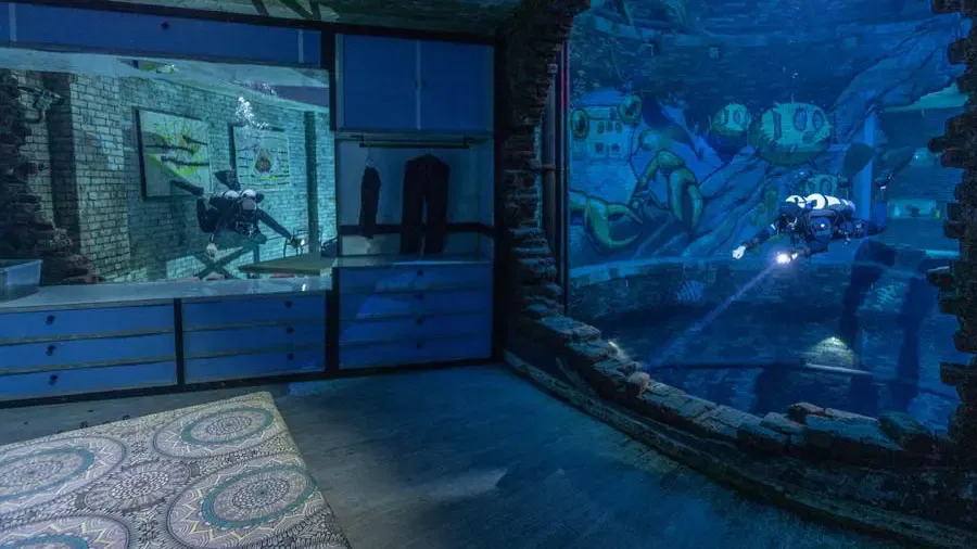 A diver swims past a submerged room with blue dressers, art on the brick walls, and a patterned bed in the foreground, near a large mural at Deep Dive Dubai.