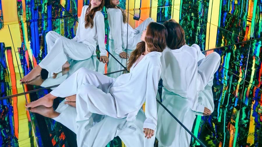 A visitor dressed in white seated on a mirrored floor, reflections multiplying against vivid yellow, green and blue striped walls at AYA Universe Dubai