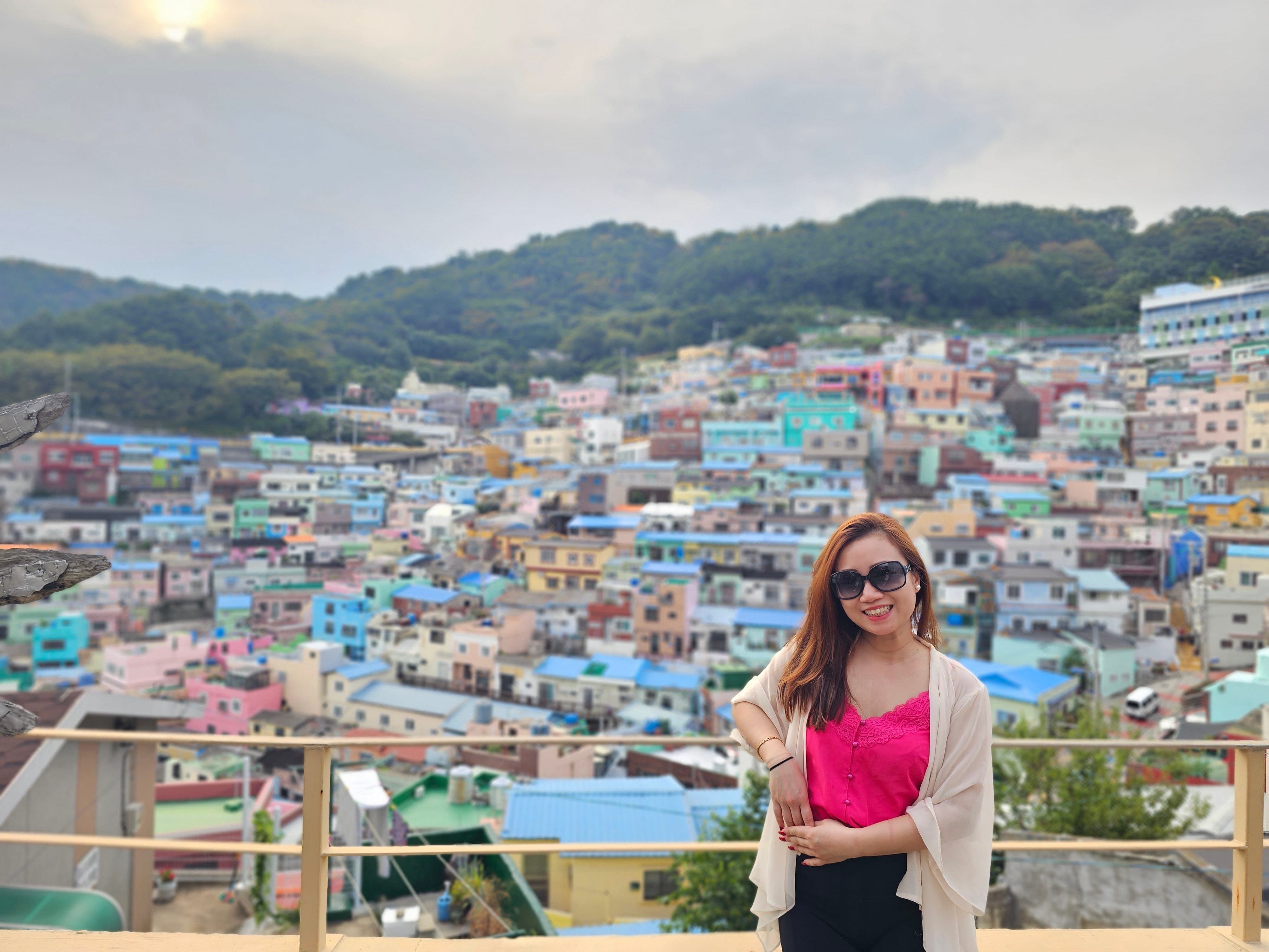 A woman in a pink top poses in front of the colorful hillside houses of Gamcheon Culture Village in Busan.