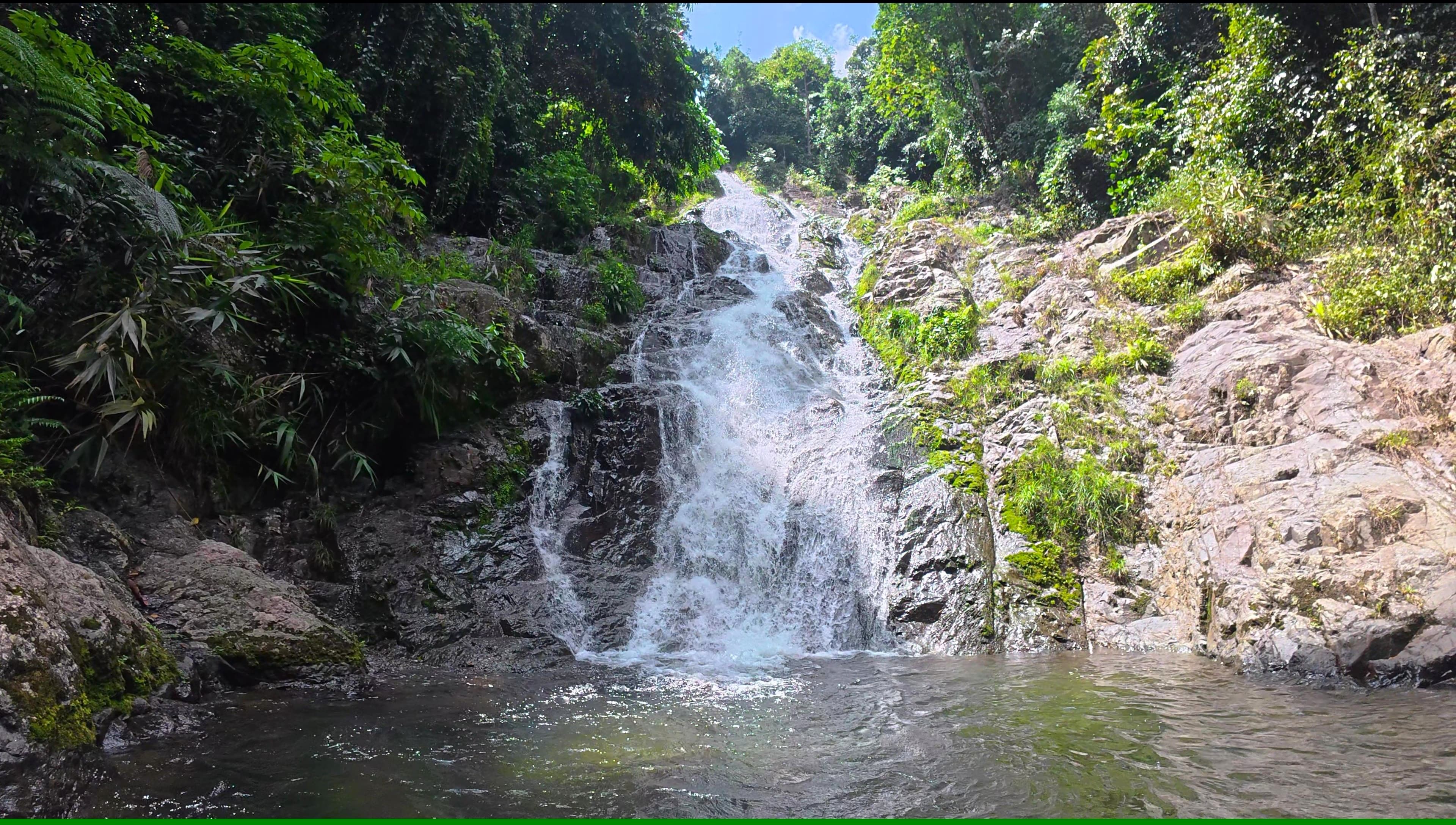 The beautiful Jeram Perlus waterfall cascades down a rocky, moss-covered slope into a clear pool, surrounded by lush green rainforest. 