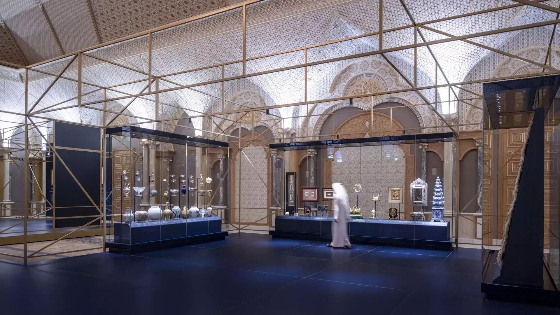 A visitor walks past glass display cases filled with artifacts in a vaulted gallery with geometric gold framing at Qasr Al Watan, Royal Palace.