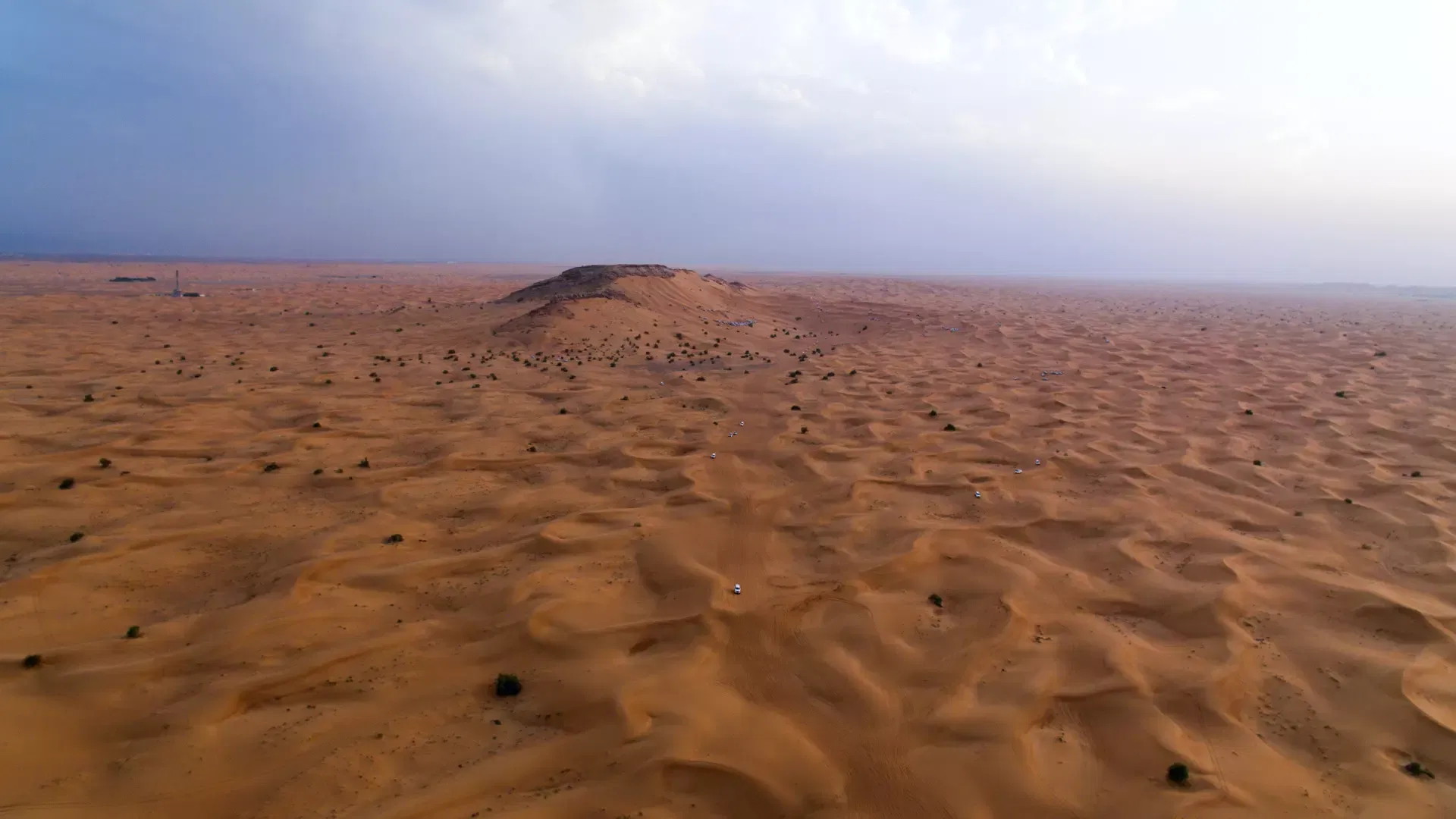 An aerial wide shot captures an expansive desert landscape of rolling sand dunes and a central rocky hill under an overcast sky at Desert Safari + Al Faqaa Camp.