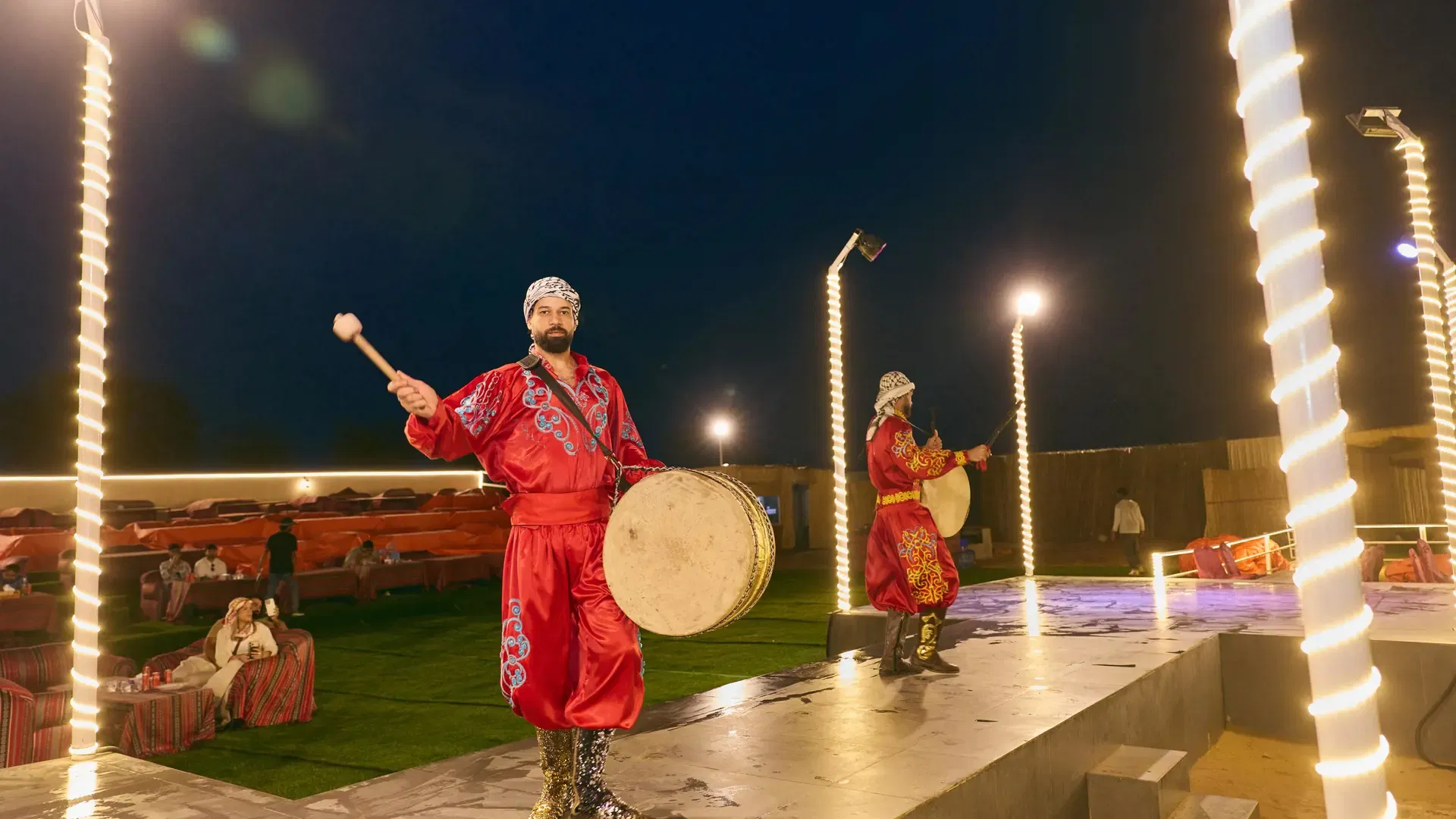 Two male musicians in traditional red attire perform on an illuminated outdoor stage at Desert Safari + Al Faqaa Camp.