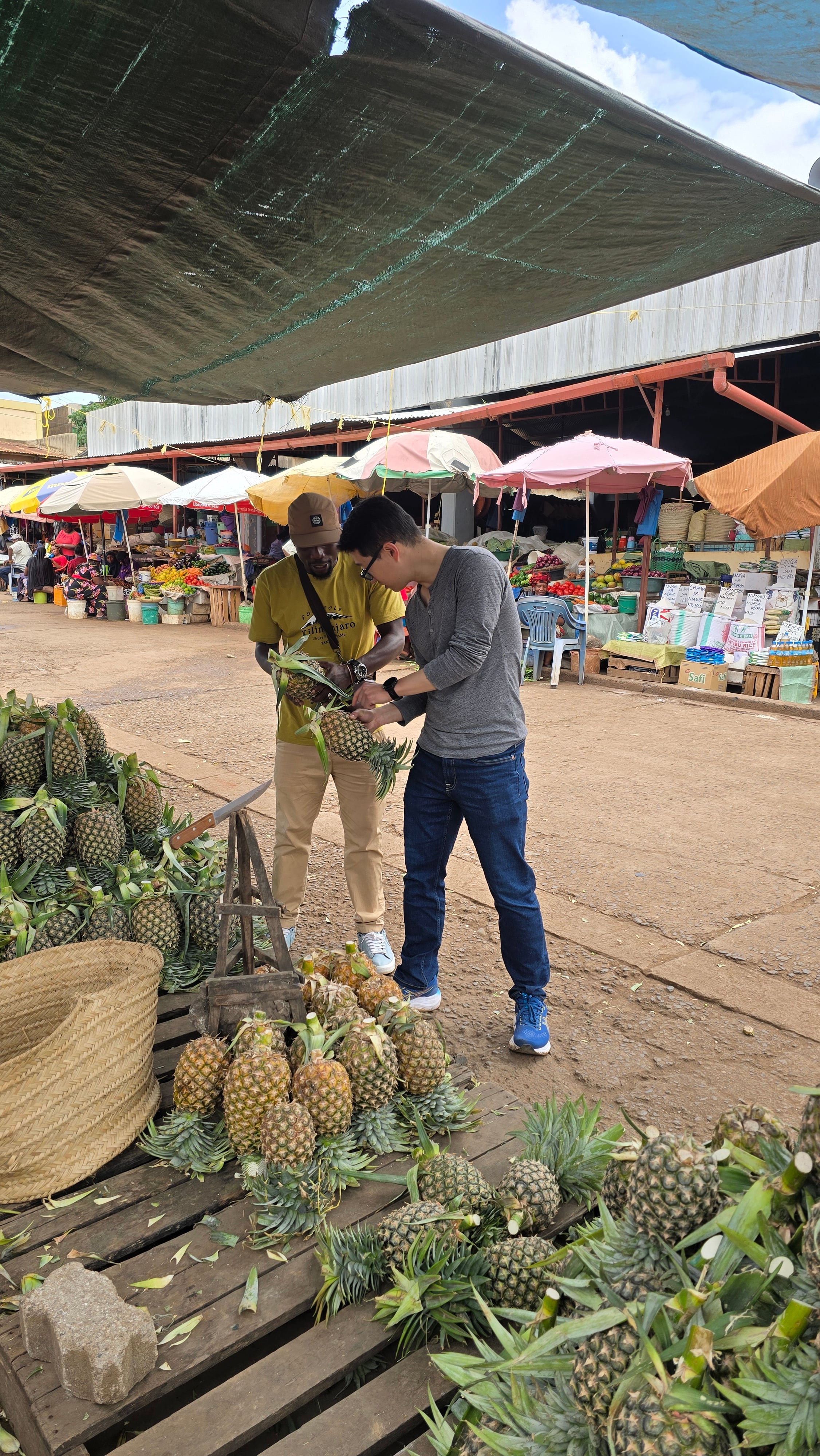 A guide and a visitor looking at fresh pineapples and fruit at an open-air market in Moshi.