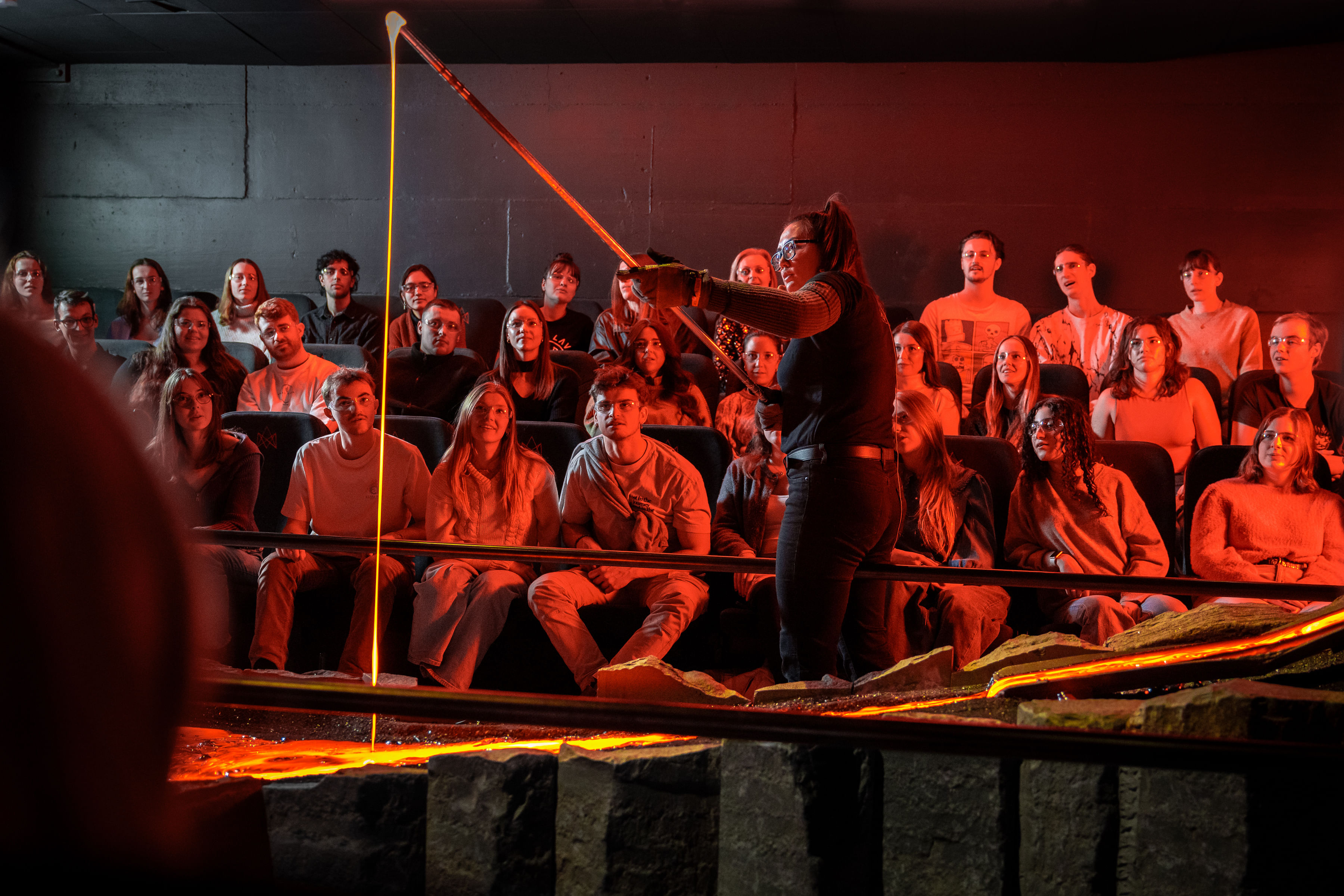 A close-up of a lava stream pouring from a tilted metal container under intense orange lighting with a person standing nearby.
