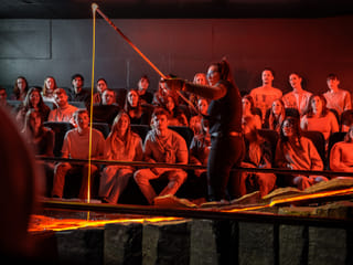 A close-up of a lava stream pouring from a tilted metal container under intense orange lighting with a person standing nearby.