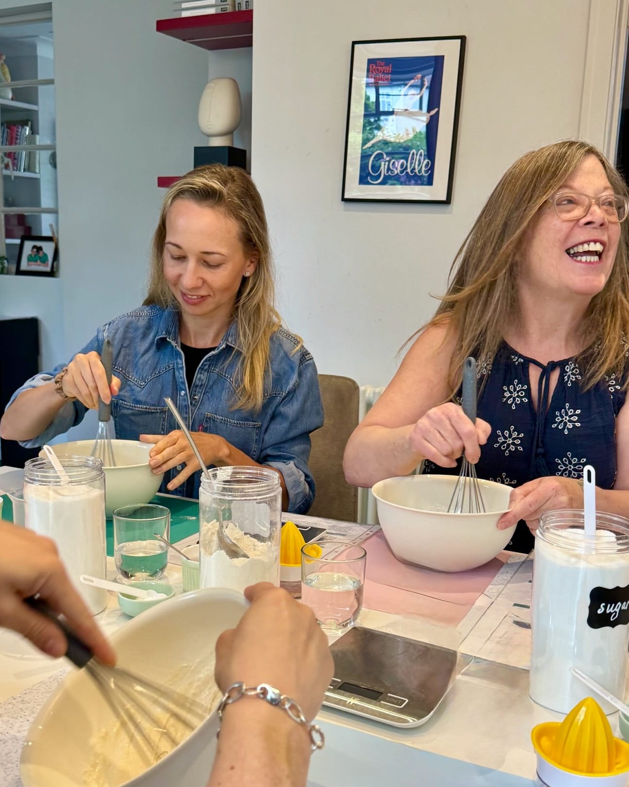 Two ladies sitting at the table whisking bowls full of crumpet batter