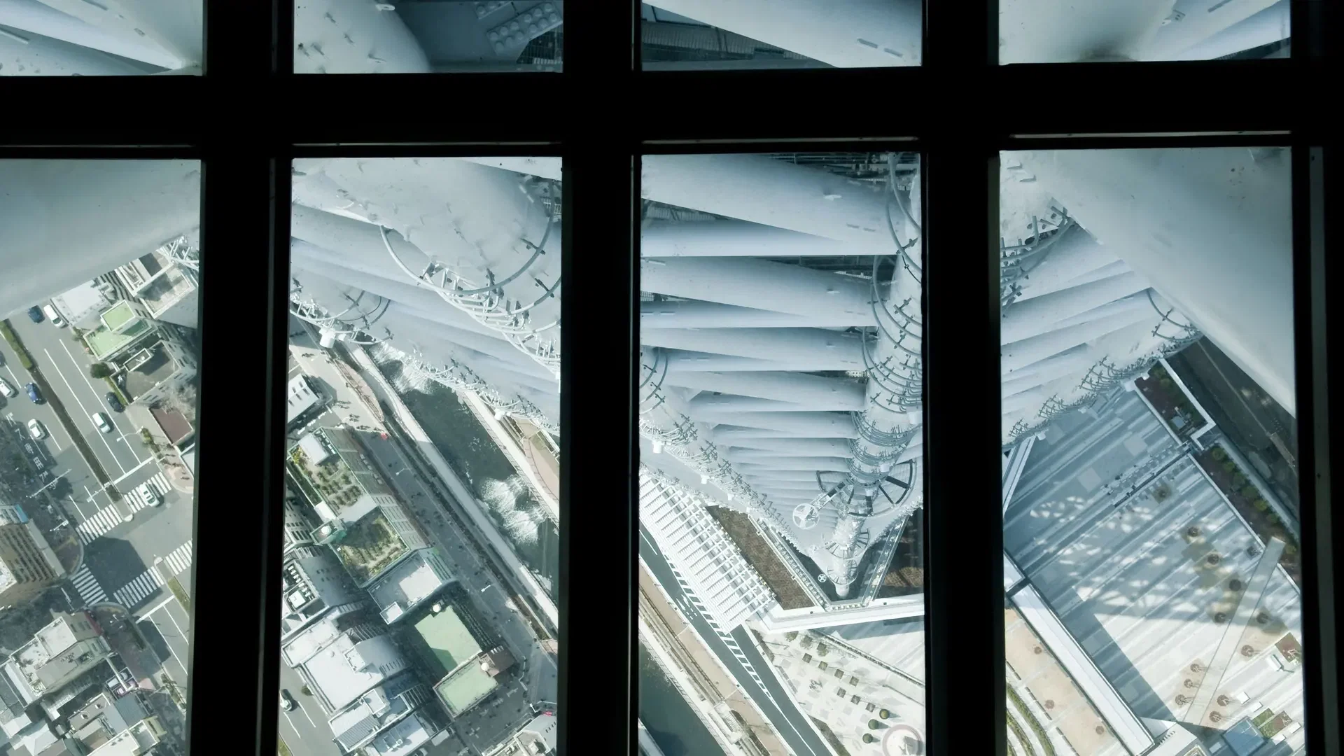 A straight-down view through window panes shows streets, buildings, and the white geometric beams of a tall tower at COMBO: Tokyo Skytree + teamLab Planets TOKYO Tickets.