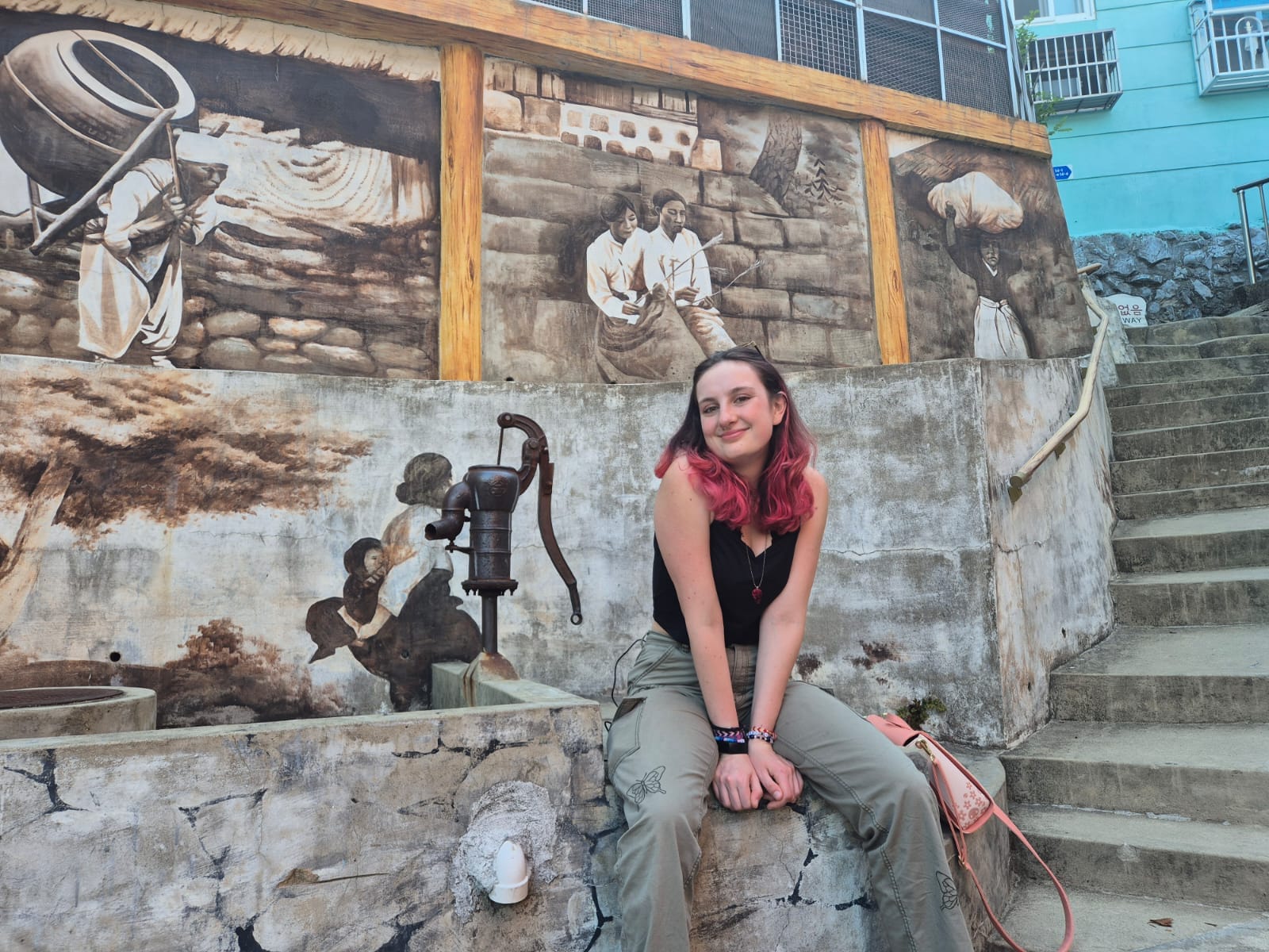 A woman smiles while sitting by the Cheondeoksu well mural in Gamcheon Culture Village, surrounded by painted stone walls.