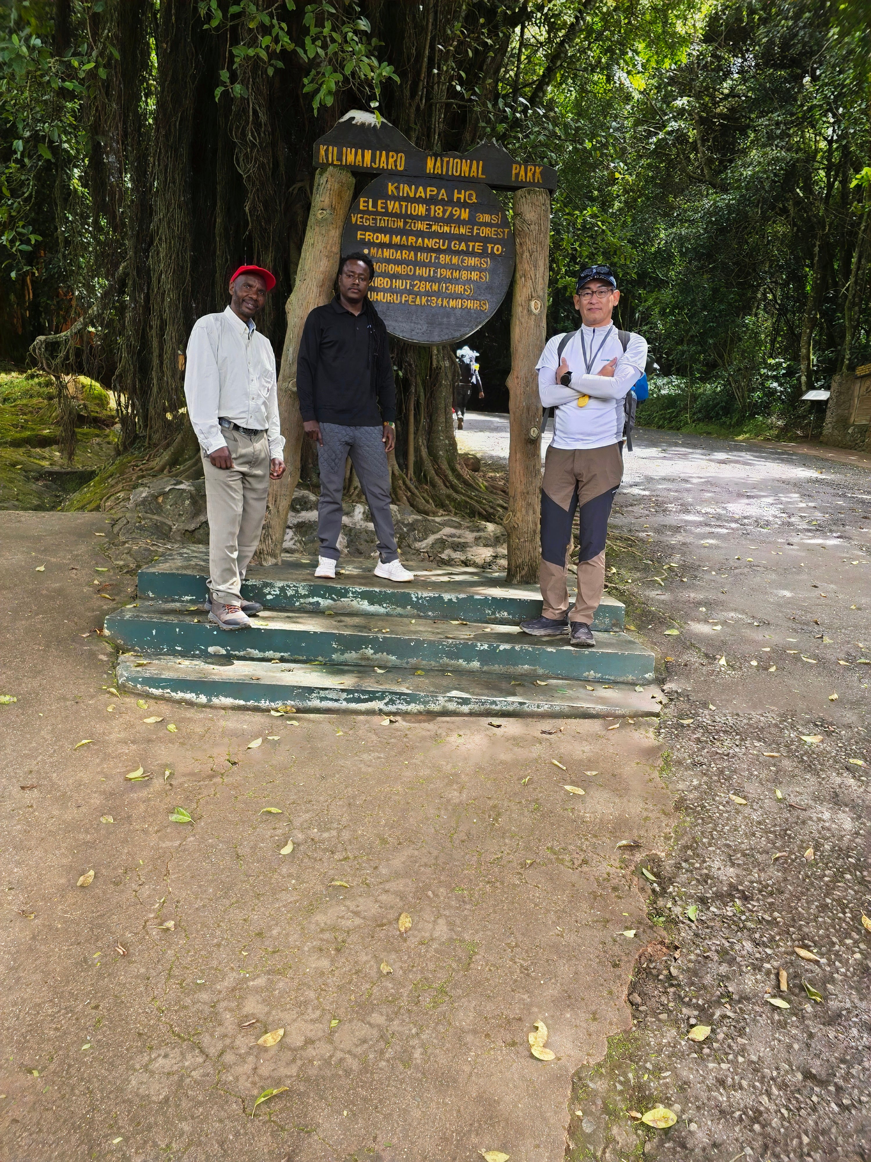 Three men standing outdoors on a dirt path near lush forest trees at the entrance of Mount Kilimanjaro National Park.