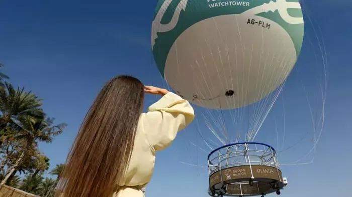 A woman with long brown hair looks up at a large tethered gas balloon at The Dubai Balloon at Atlantis.