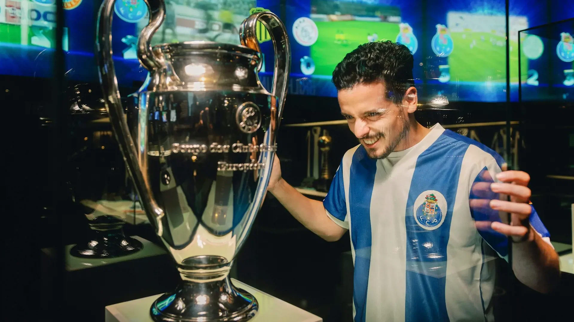 A smiling man wearing a blue-and-white jersey poses next to a large trophy encased in glass with digital screens behind him at FC Porto Museum & Dragão Stadium ticket.