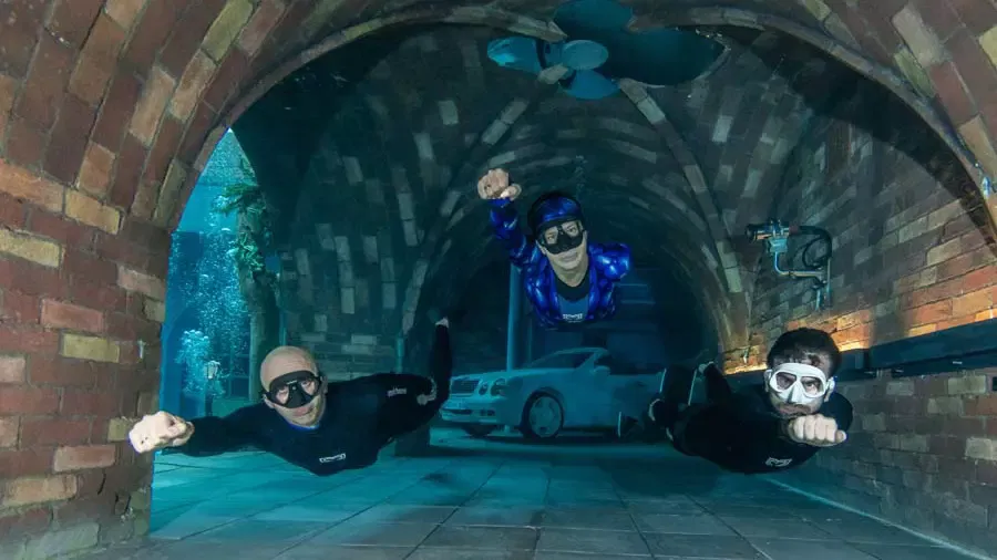 Three freedivers swim in a flying pose through a submerged brick archway with a car in the background at Deep Dive Dubai.