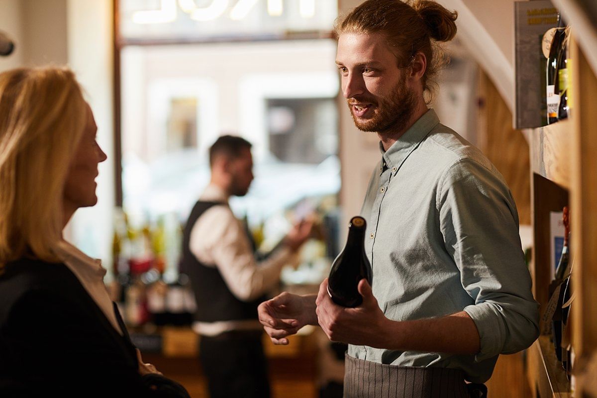 A friendly wine expert discussing a bottle of wine with a guest in a cozy tasting room, with a bartender preparing drinks in the background.