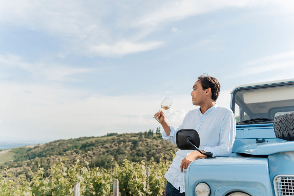 A man is enjoying a glass of wine in the vineyards while leaning on the side of a vintage Land Rover Defender