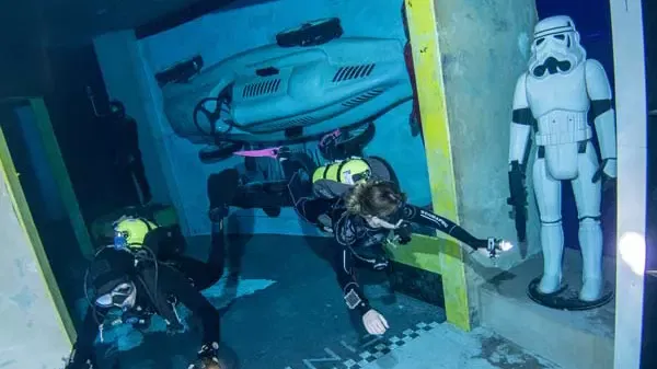 A scuba diver swims past a white armored figurine and a submerged car mounted on a wall at Deep Dive Dubai.
