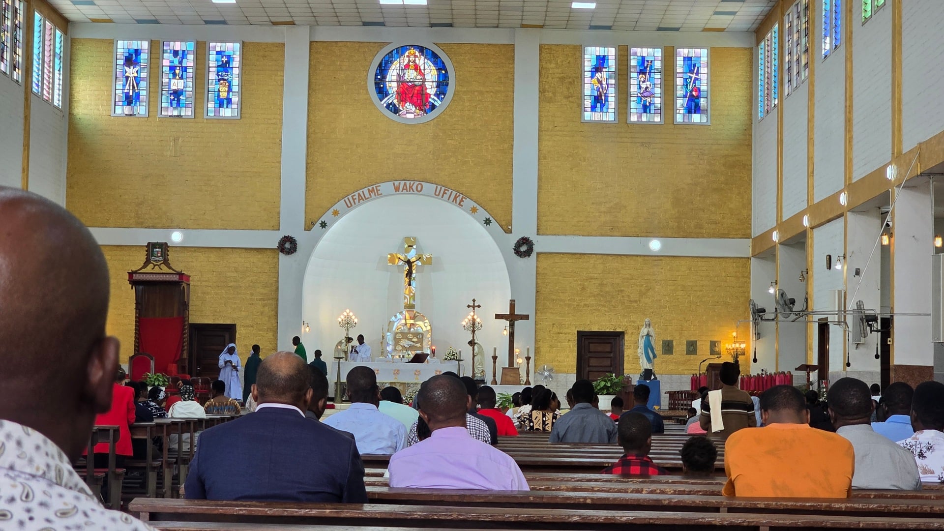A wide view of people seated inside Christ the King Cathedral facing the altar and stained-glass windows.