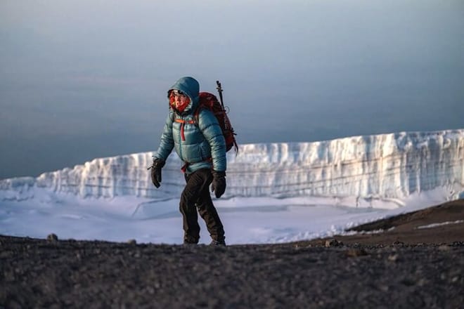 A hiker in a blue puffer jacket walking on a dark volcanic ridge with a massive white ice glacier in the background.