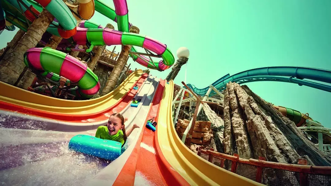 A smiling young girl rides a blue mat down a multi-lane rainbow water slide with other visitors visible on adjacent slides at Yas Waterworld, Abu Dhabi.